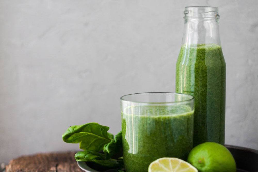 Glass and bottle of green smoothie sitting next to spinach and limes on table